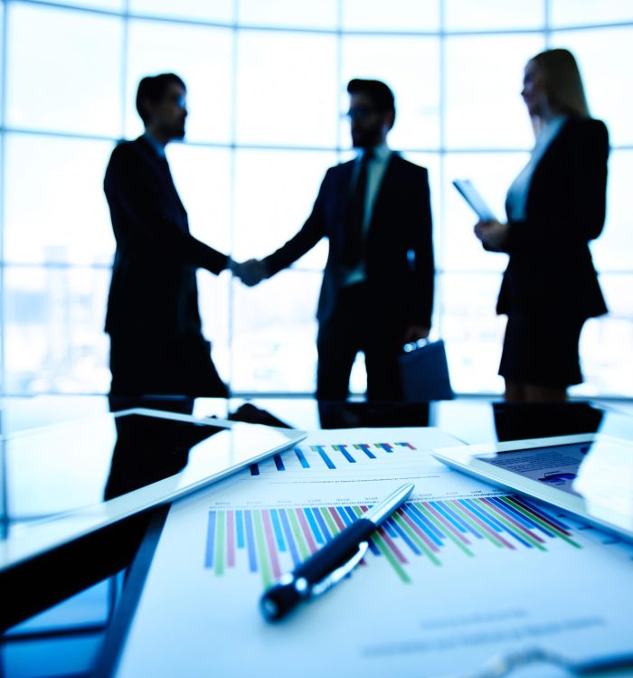 Technological devices, financial document with pen, glass of water at workplace on background of three business partners striking deal